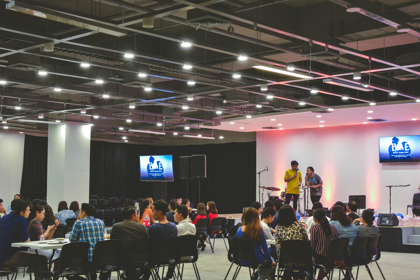 A business conference with groups of people sitting around tables with a man and a woman presenting on a stage